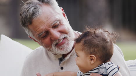 senior hispanic man sitting in the garden with his baby grandson on his knee, talking to him, head and shoulders close up