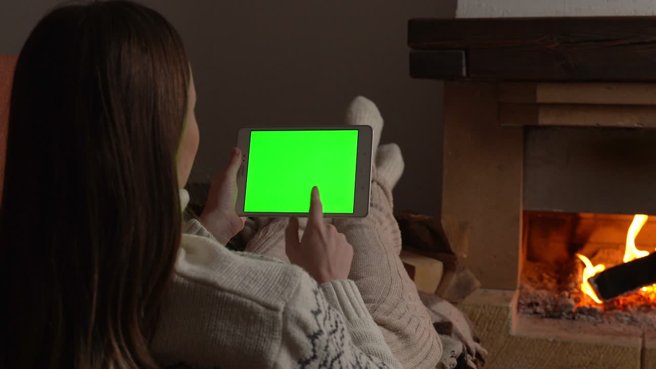 donna sdraiata sul divano di fronte al camino tenendo in mano lo schermo verde del tablet, guardando lo schermo chiave chroma vicino al camino