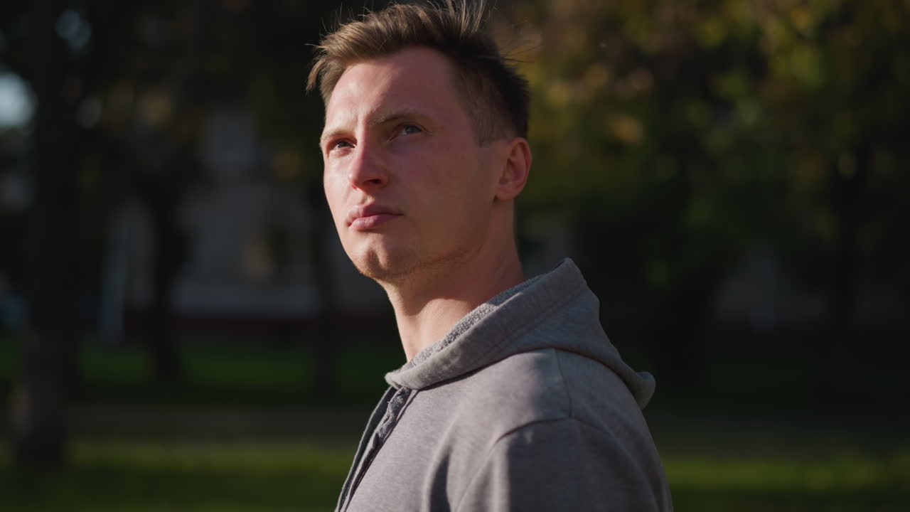 Young man with short dark brown hair wearing light gray hoodie, serious and contemplative expression while looking into distance, with blurred green and yellow-brown foliage in background