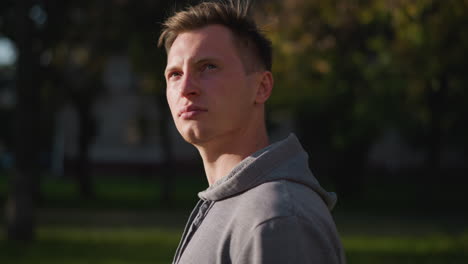 young man with short dark brown hair wearing light gray hoodie, serious and contemplative expression while looking into distance, with blurred green and yellow-brown foliage in background