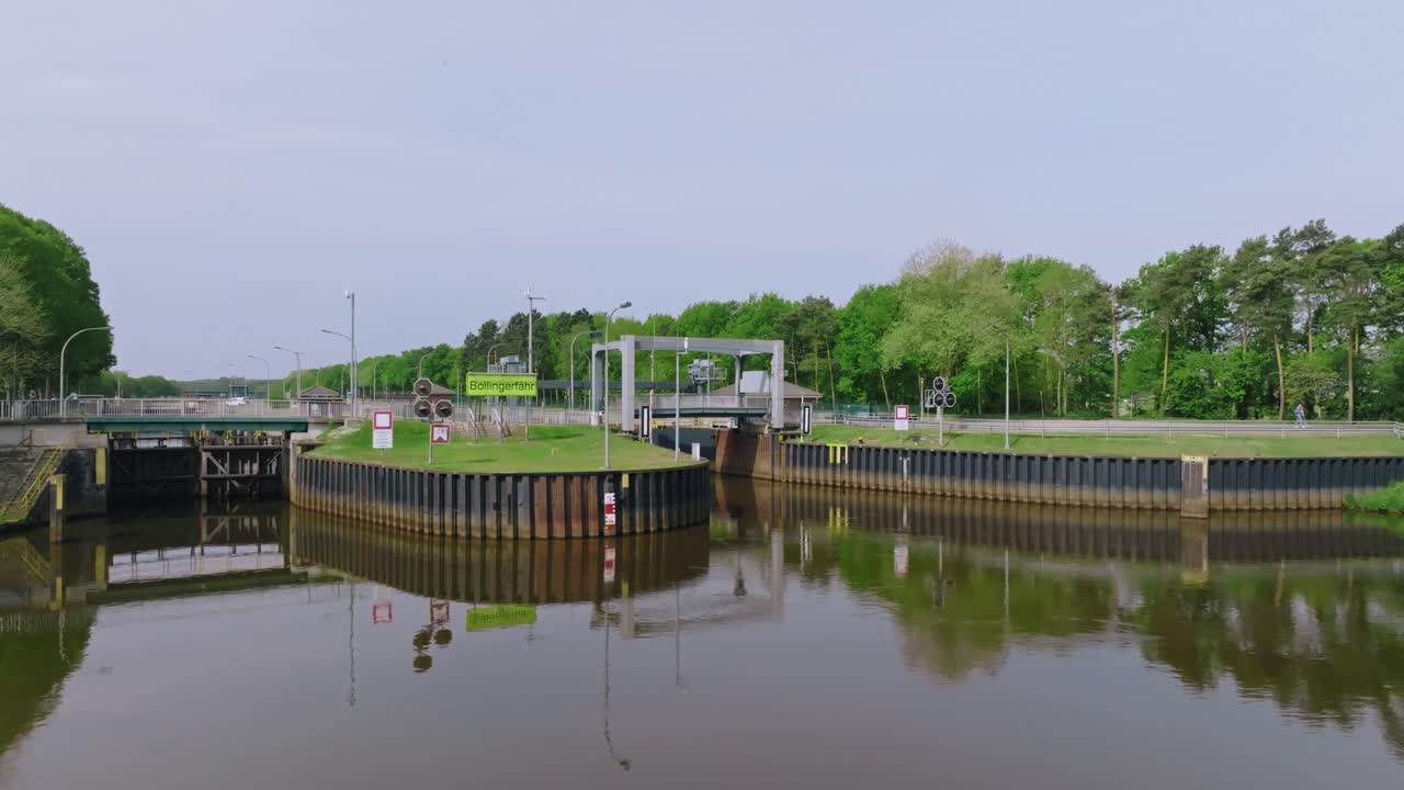 Pan right across the entrance of a water lock surrounded by green fields and road access near the forest.