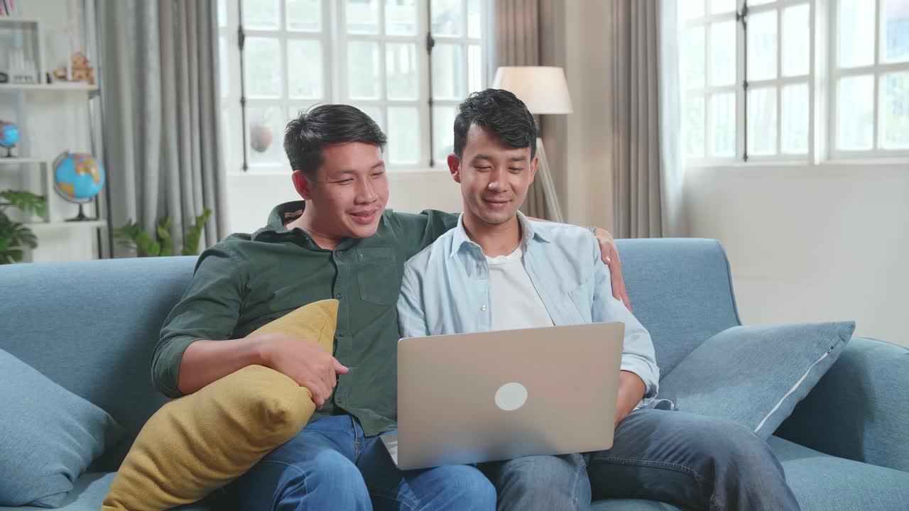 Portrait Of Asian Gentle Gay Couple Using Laptop Computer, While Sitting On A Couch In Cozy Stylish Apartment. Males Looking Each Other