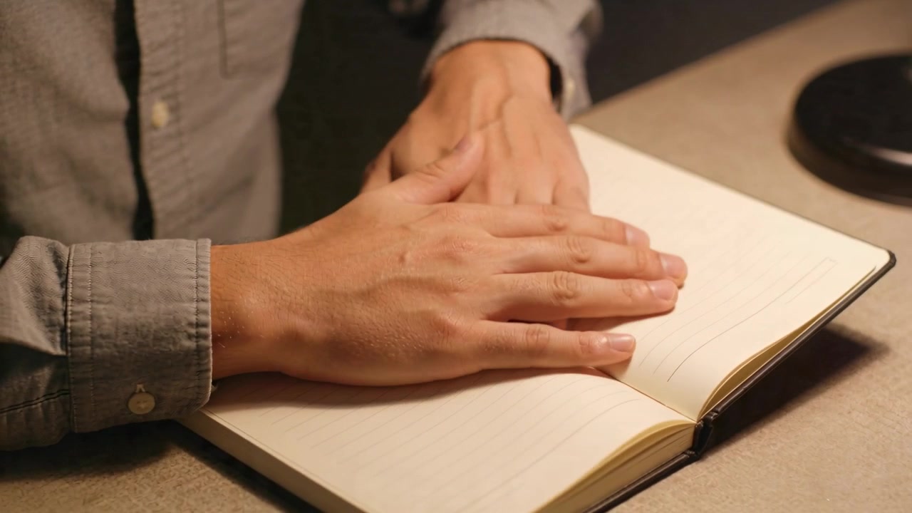 Mani maschili alzano la mano destra, sfogliano le pagine e premono un quaderno sulla scrivania vicino a una lampada, con polsino e anello visibili.