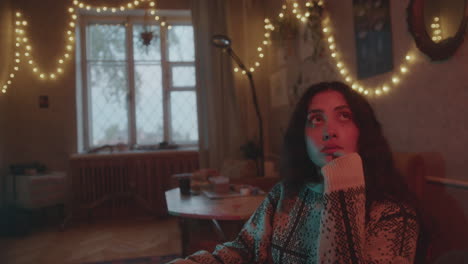 thoughtful girl sitting alone in cozy living room with string lights