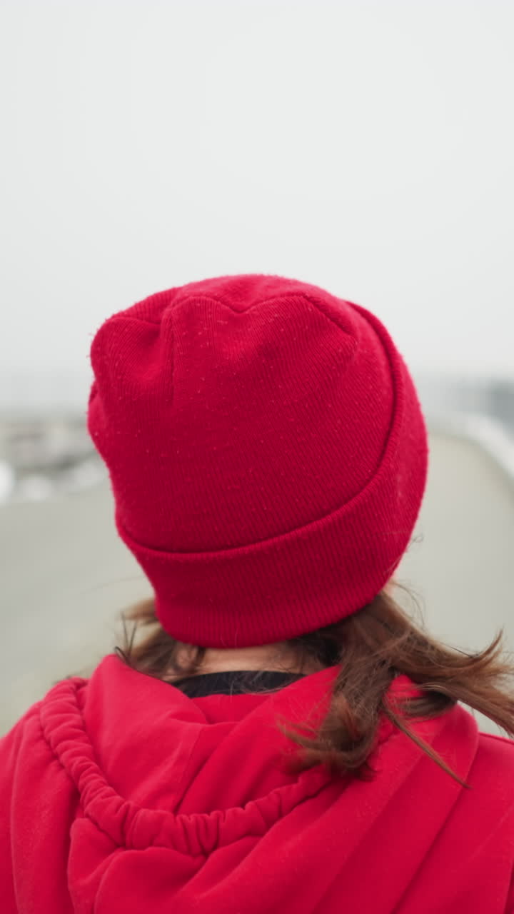 close up back view of woman jogging in red beanie and jacket along snowy pathway near iron railing benches in foggy winter park with distant bridge light poles snow covered trees visible breath cold