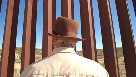 curious man with fedora hat looks through border fence dividing united states and mexico