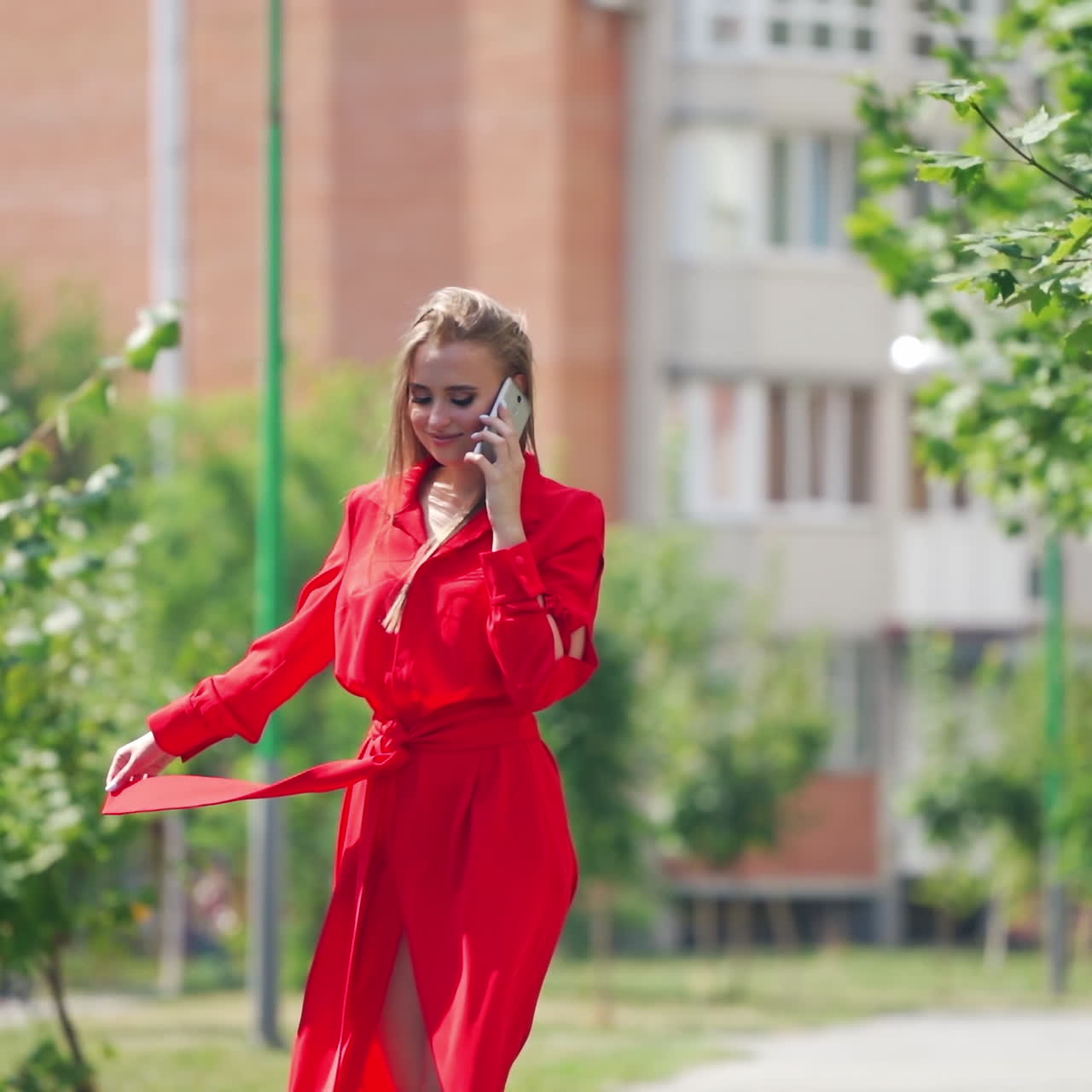 Sexy woman in red dress outdoors. Young lady walking slowly on the street and talking on the mobile phone. Beautiful female in passionate dress in a summertime.
