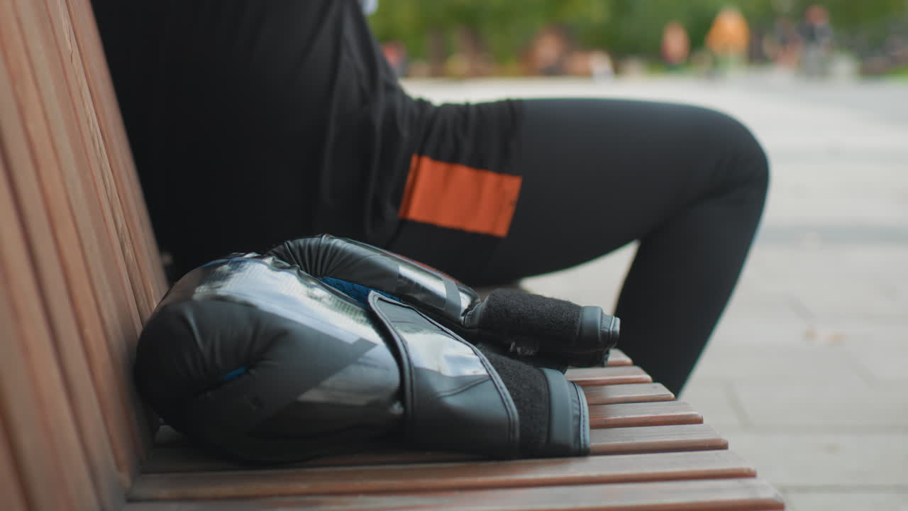 white man seated on wooden bench removing gloves and grabbing water bottle, orange shorts patch visible, stretching leg and breathing deeply, park background with distant pedestrians, relaxed recovery