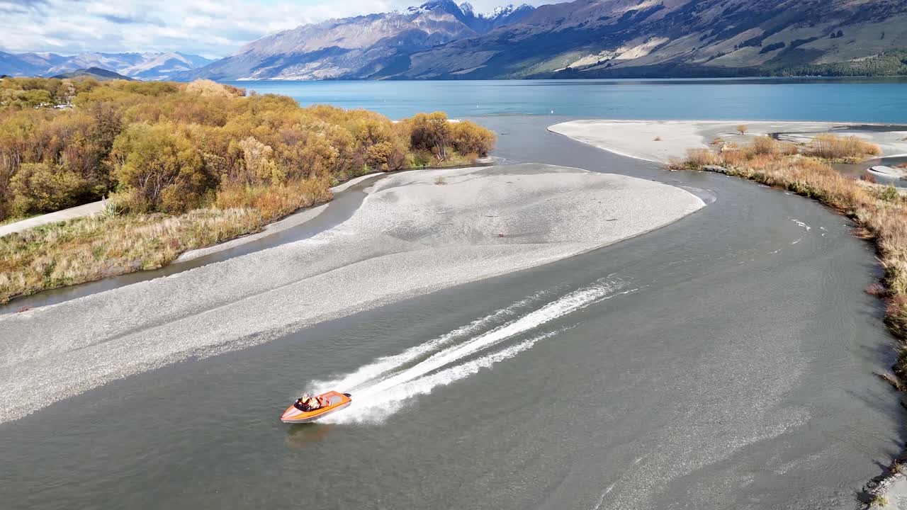 A jet boat speeds through a winding river in Glenorchy, New Zealand, surrounded by lush landscapes and distant mountains under bright daylight