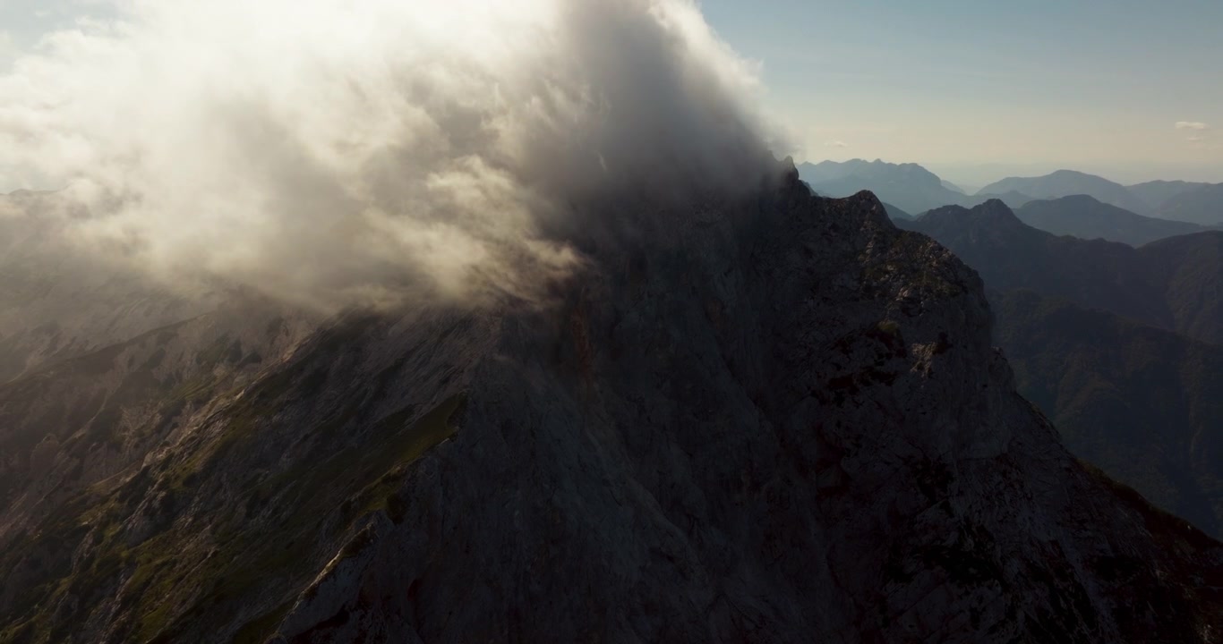 Le nuvole si muovono su una vetta montuosa frastagliata nelle Alpi di Kamnik e della Savinja in Slovenia, fluendo sulla cresta rocciosa mentre la luce del sole si fa strada.
