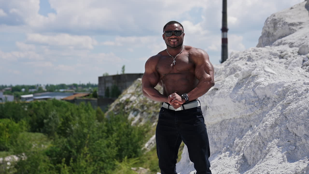 Shirtless afro american sportsman outdoors. Muscular bodybuilder with dark skin in sunglasses posing on hill on factory background.