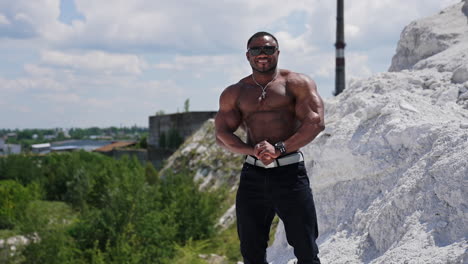 shirtless afro american sportsman outdoors. muscular bodybuilder with dark skin in sunglasses posing on hill on factory background.
