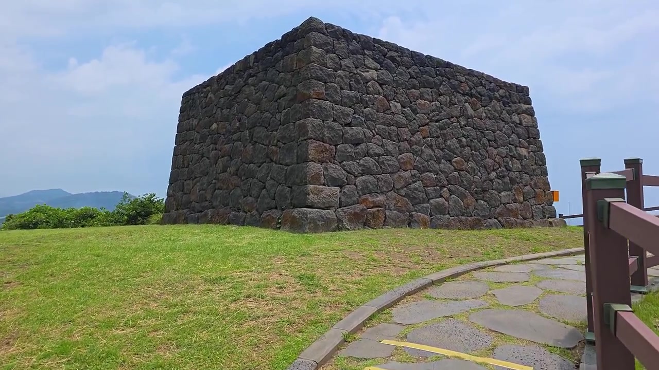 POV (punto di vista) che si avvicina al faro di segnalazione a fumo Sanbang Yeondae, una torre trapezoidale di pietre vulcaniche impilate su un tumulo erboso con una montagna sullo sfondo a Jeju, Corea del Sud.
