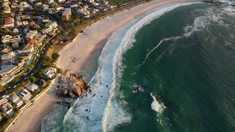 hermosa playa de arena blanca en camps bay, ciudad del cabo con olas turquesas rompiendo al atardecer, antena