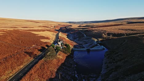 Aerial-Moorland-View-moving-past-a-farmhouse-on-a-quiet-road-with-a-river-on-Saddleworth-moor,-showing-a-wild-landscape-with-traffic,-reservoir-and-snow-lined