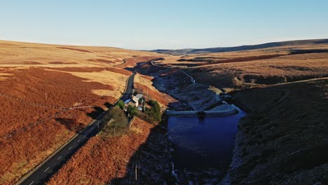 aerial moorland view moving past a farmhouse on a quiet road with a river on saddleworth moor, showing a wild landscape with traffic, reservoir and snow lined