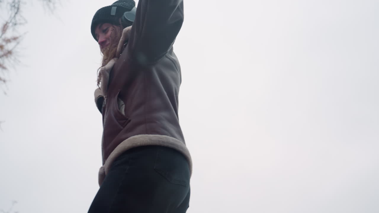 Close view of happy lady in black knit cap, brown shearling jacket with arms outstretched while walking joyfully on a cool autumn day in peaceful outdoor setting