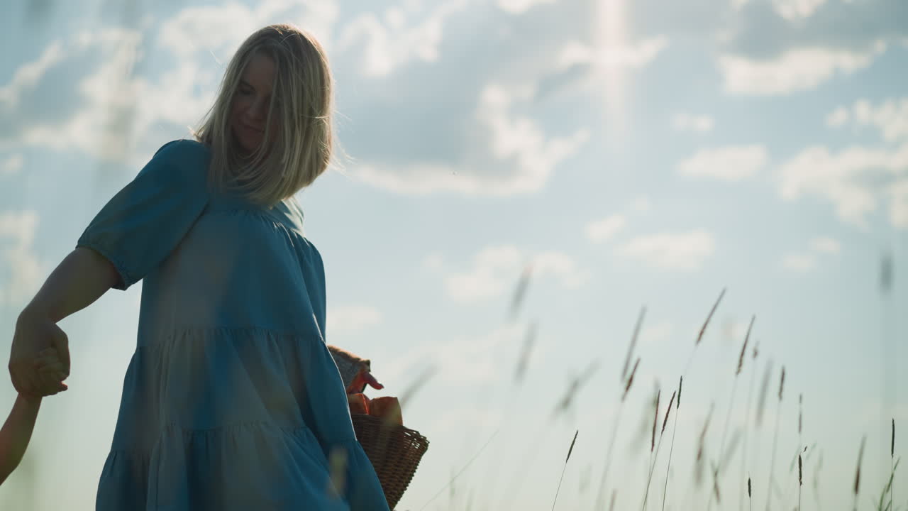 A woman in a flowing blue gown walking through a lush green grass field with her two young sons, both wearing white shirts. She carries a basket filled with fruits, under a clear sky with soft clouds