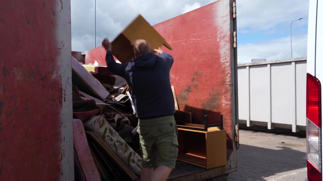 Man in hoodie and shorts hurls and stacks old furniture units into a red metal skip at a public dump site.