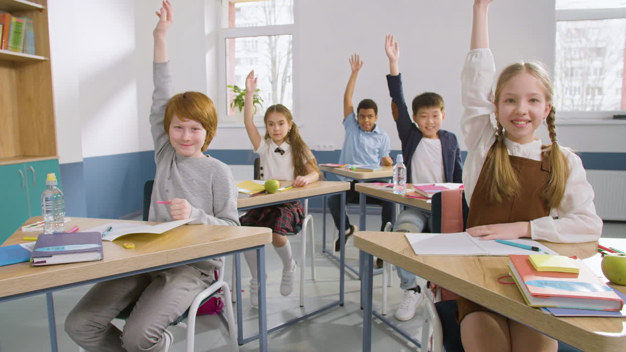 многонациональная группа студентов сидит за партами в классе английского языка и пишет в тетрадях, затем поднимает руки, чтобы ответить на вопрос учителя 1