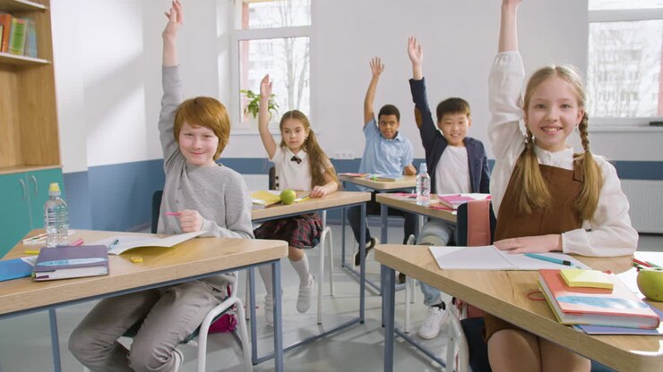 Multiethnic Group Of Students Sitting At Desks In English Classroom Writting In Their Notebooks, Then Raise Their Arms To Answer The Teacher's Question 1