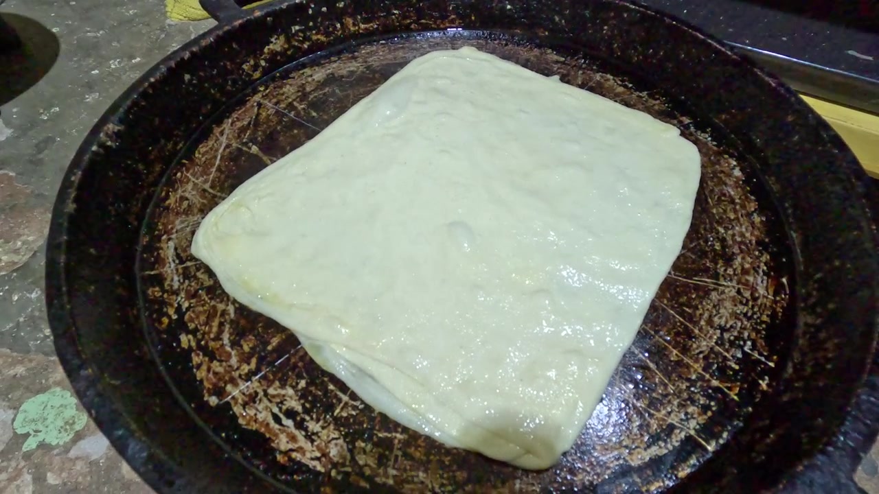 Msemen marocchino tradizionale che cuoce su una padella di ferro stagionata. L'impasto sfogliato piegato e unto d'olio inizia a dorarsi, evidenziando l'autentica preparazione di pane piatto fatto a mano utilizzando tecniche culinarie tradizionali