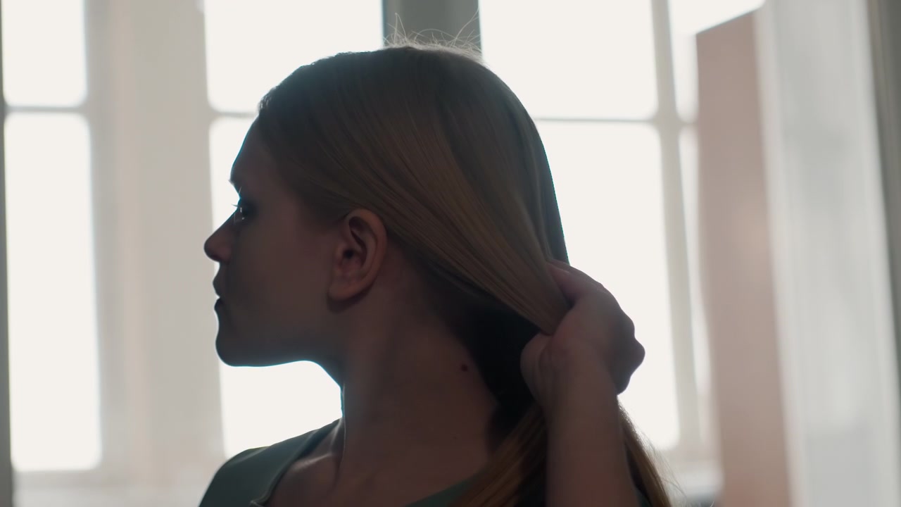 Donna caucasica allo specchio della finestra della camera da letto che guarda di lato e si liscia i capelli, silhouette in controluce soffusa ed espressione contemplativa. Ritratto intimo che mostra il profilo, sottili gesti di toelettatura.