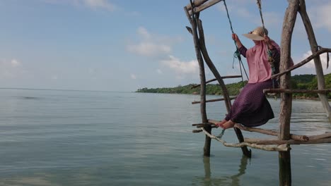 woman in summer hat swinging on wooden swing over shallow and calm water at karimun jawa island with camera tilt up and beautiful blue sky background