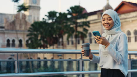 young muslim woman using smartphone and holding coffee cup in city