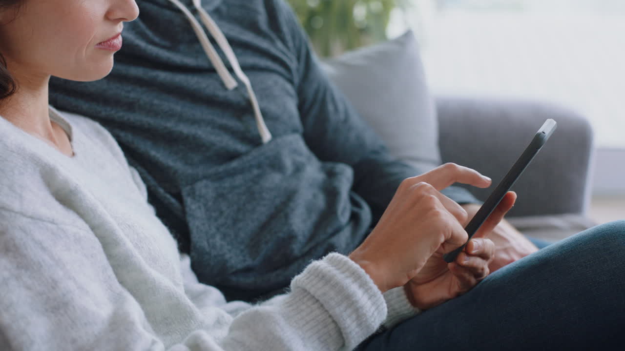 행복한 커플이 집에서 스마트폰을 사용하여 함께 휴식을 취하고 온라인 쇼핑을 즐기고 인터넷을 탐색하고 소파에서 휴식을 취하고 있습니다.