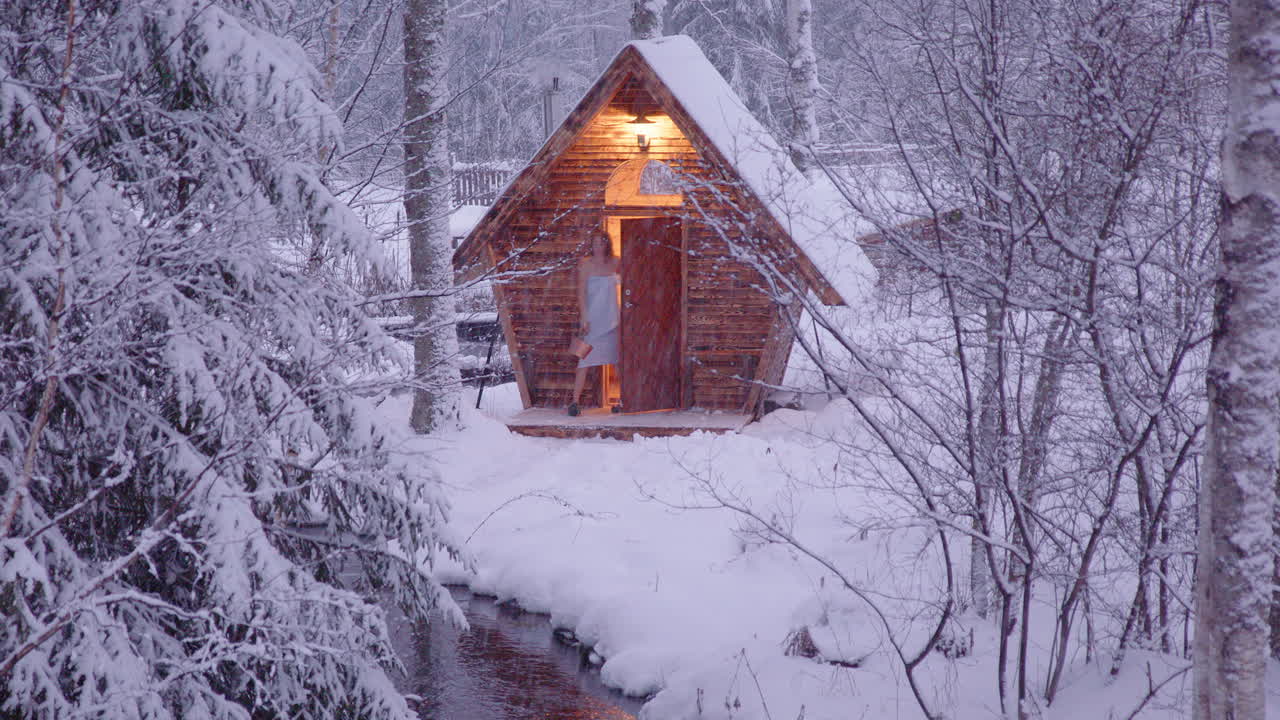 Woman in towel exits wooden sauna in snowy woods to collect water from stream
