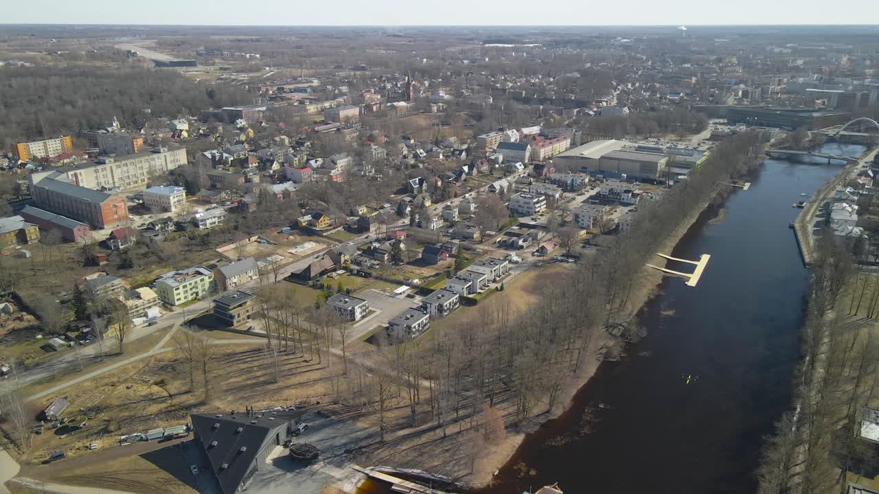 Aerial drone footage panning over at Tartu city in Estonia where small apartment houses and complexes are visible around Tartu Emajõgi during a sunny spring day. Boats visible in the cold dark river.