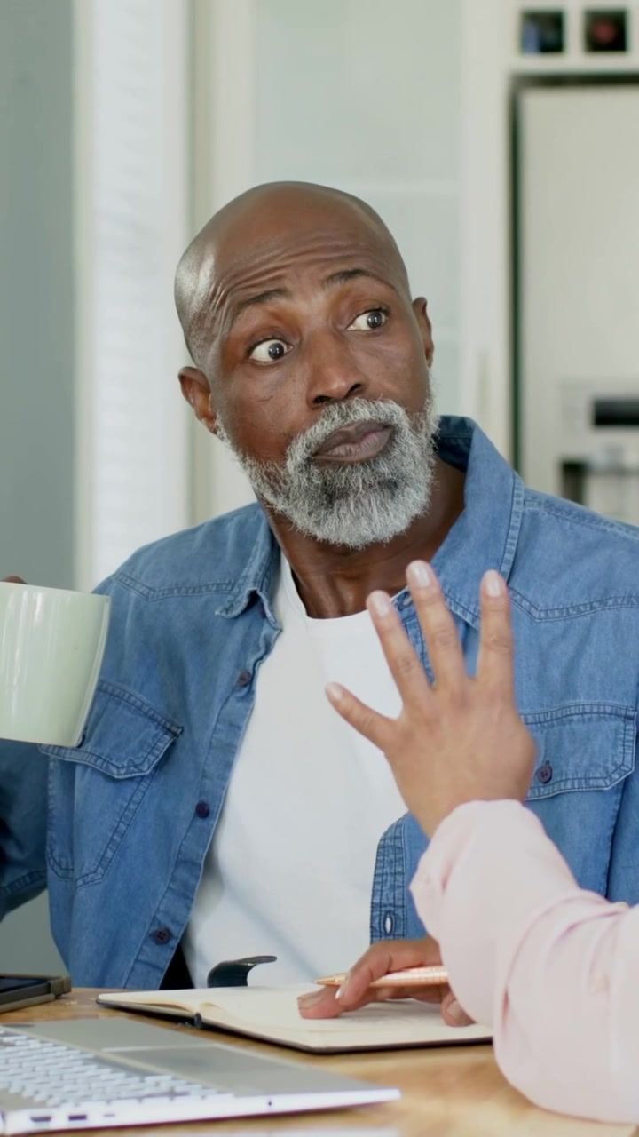 Video verticale: Uomo in jeans che sorseggia da una tazza di menta e reagisce a un collega che gesticola nell'ufficio di casa, con un notebook.