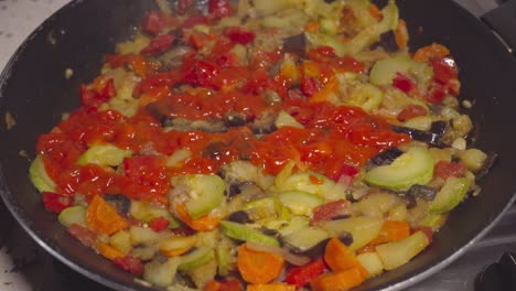 agregando pasta de tomate al pisto español y revolviendo, primer plano a mano y sartén