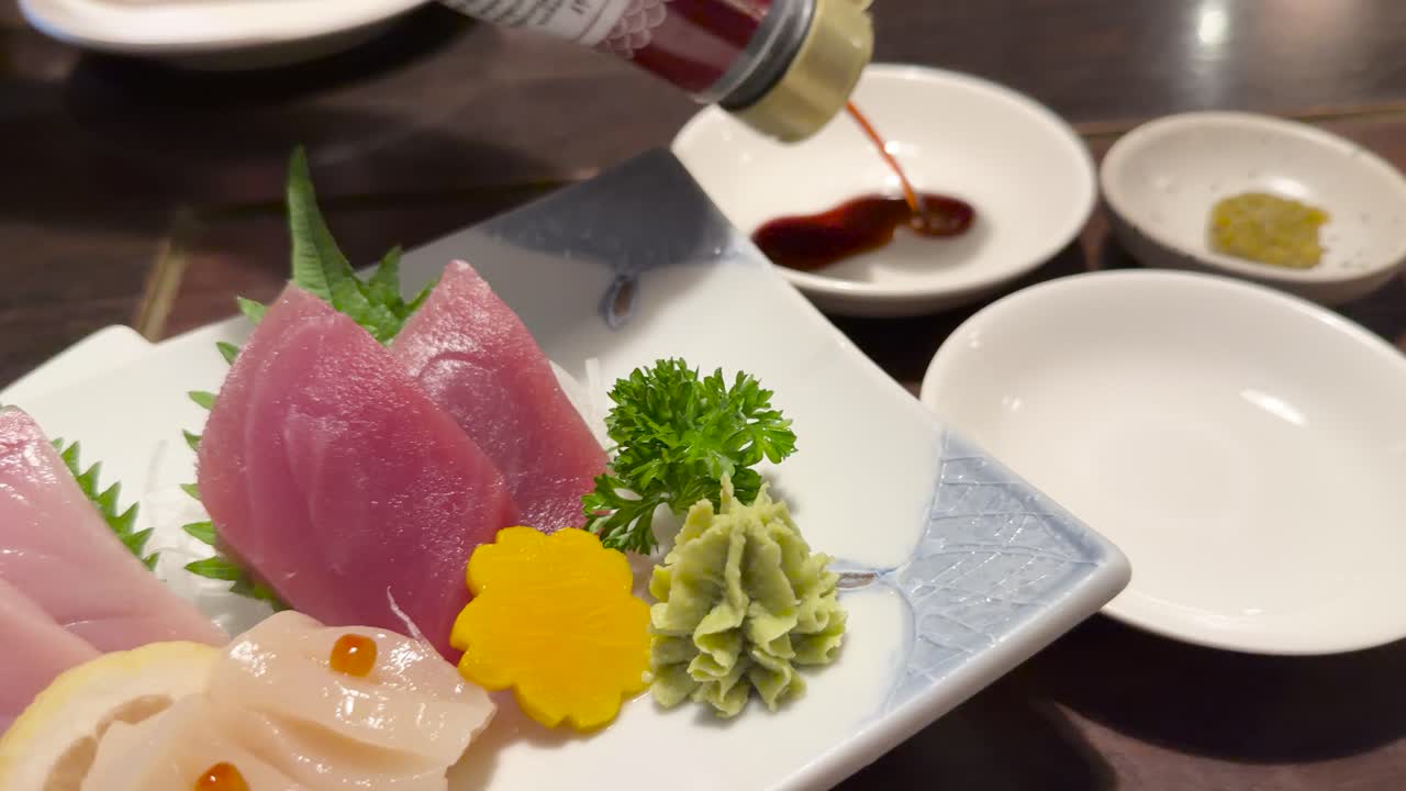 Hand pours soy sauce into dipping dish beside assorted sashimi in warmly lit Japanese restaurant