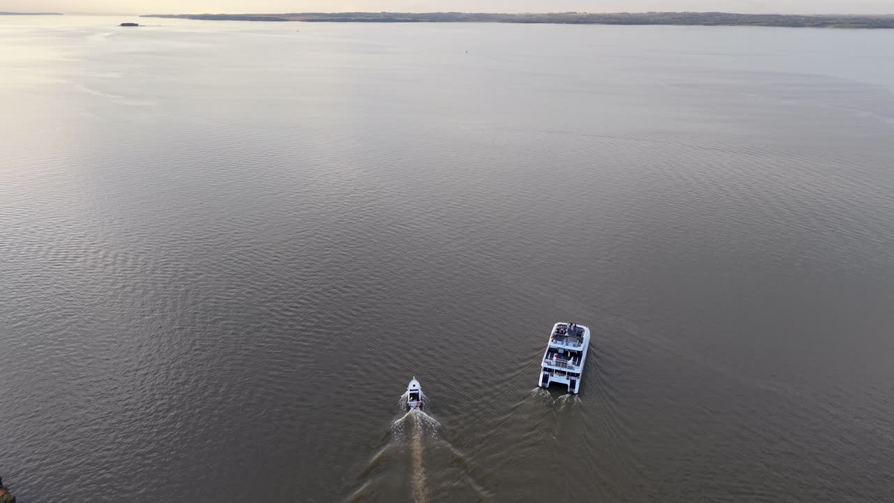 Accelerating aerial drone shot approaching a white catamaran and escort boat sailing away from the dock into the open river on a cloudy afternoon