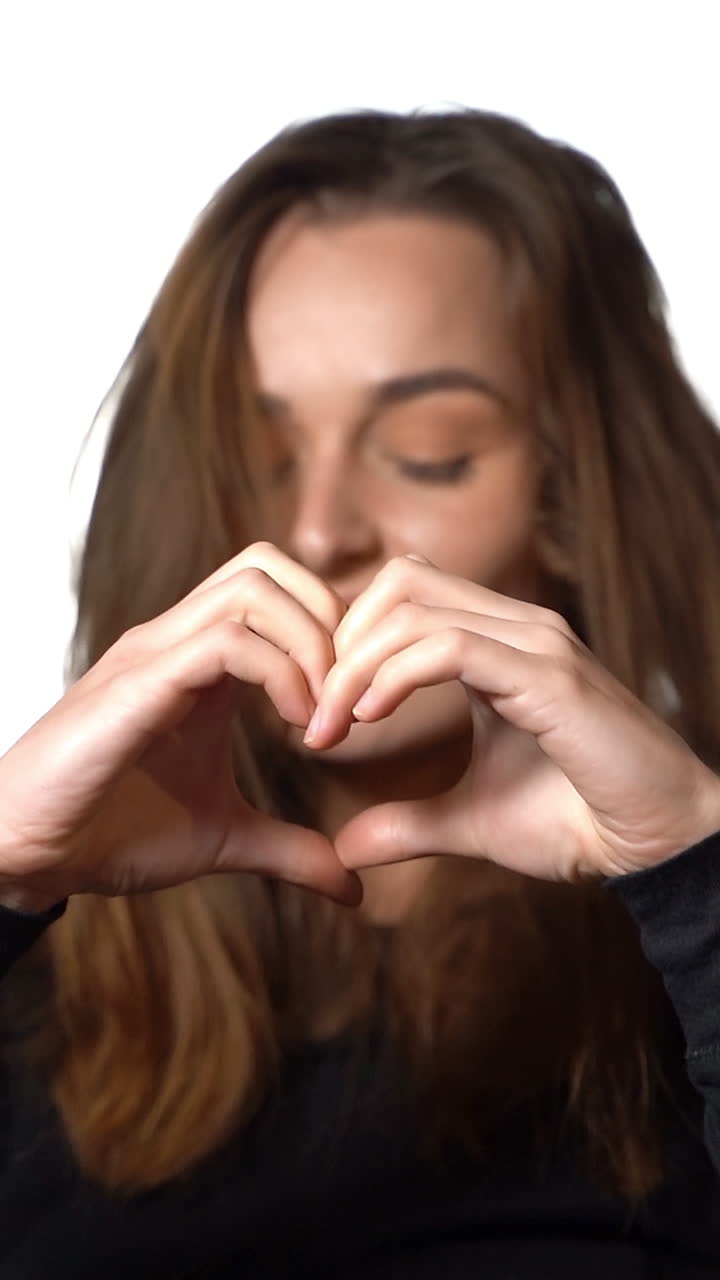 Woman is portraying heart by her hands before her face on a white background in the studio. Close-up Vertical video