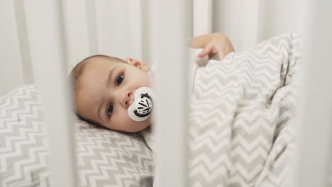 portrait of a baby girl falling asleep in a crib with a pacifier in her mouth