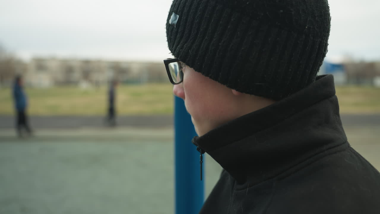 Close-up of a young boy standing near a blue pole, looking thoughtful as he gazes away toward the field, with a blurred view of someone standing by the track and people passing by