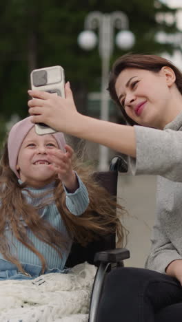 toothless girl makes videos with mom waving hello in camera of smartphone. smiling woman and child in wheelchair make content for social media in park