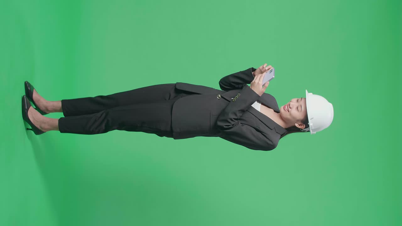 Full Body Side View Of Smiling Asian Female Engineer Looking Around And Shaking Her Head While Using Smartphone In The Green Screen Studio