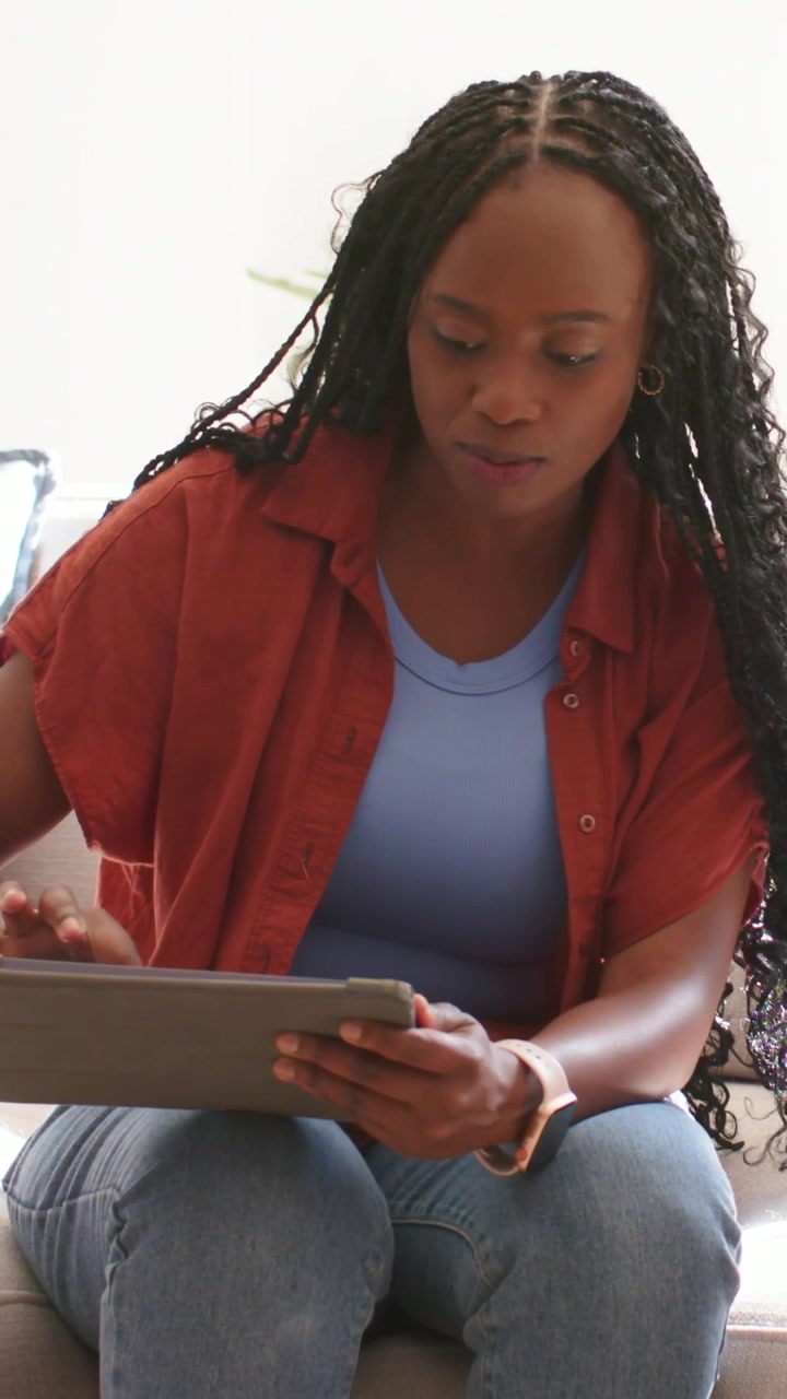 Vertical video: Tapping tablet after glancing right, woman in rust top browsing messages on sofa