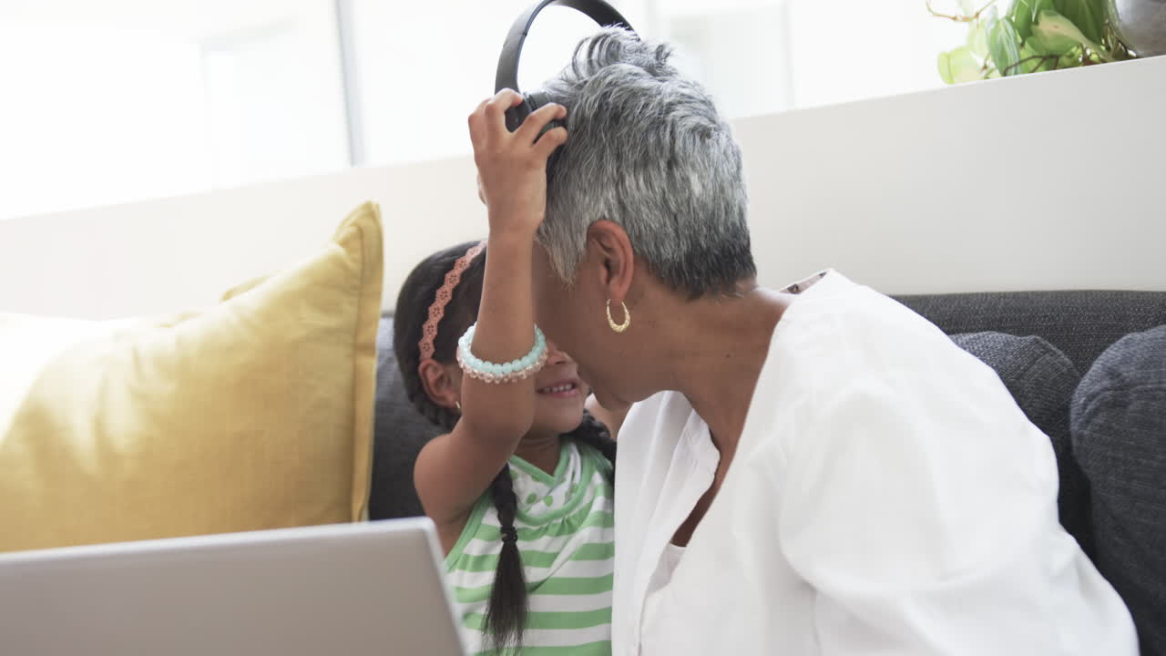 Biracial girl shares headphones with her grandmother, both smiling in a home setting