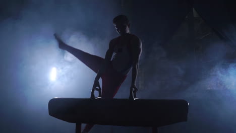 male gymnast performs exercises from the olympic program on pommel horse performing spins and rolls in slow motion getting ready for the olympics by training hard in the dark room around the smoke and the counter light