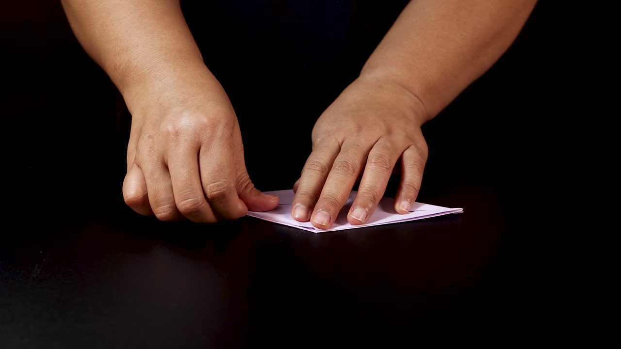 Adult hands carefully fold white paper on black table under soft, even lighting, overhead view