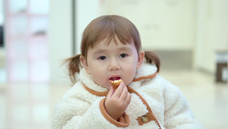 cute 3 years old toddler in ponytails and fluffy coat enjoys eating bread inside a shopping mall with blurred view of people walking behind