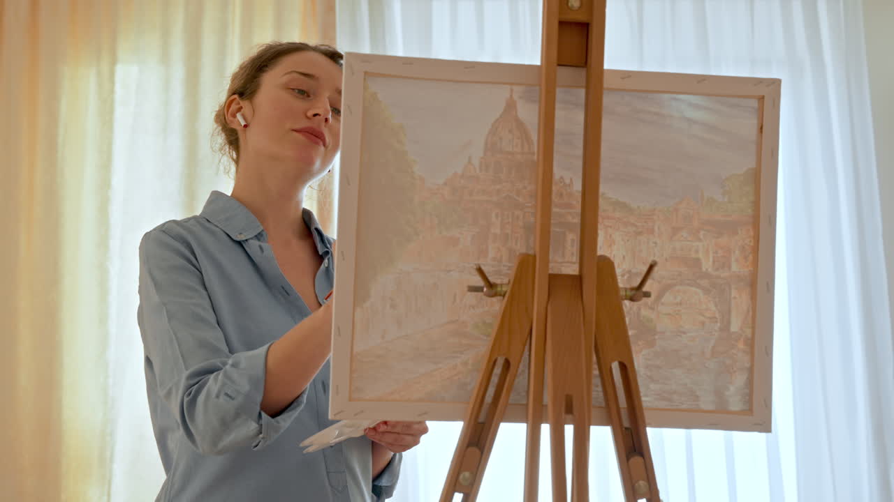 Caucasian woman painting a picture with a palette in her hands and listening music with earphones indoors
