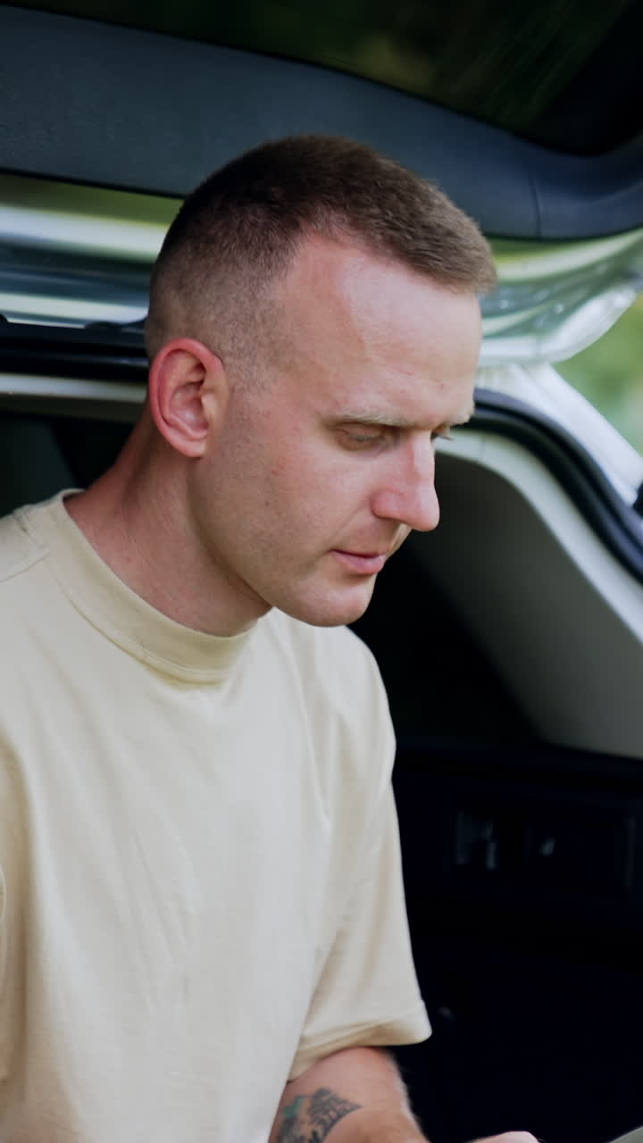 Focused male freelancer looks at the screen of his laptop. Man works remote sitting in the car trunk in forest. Vertical video