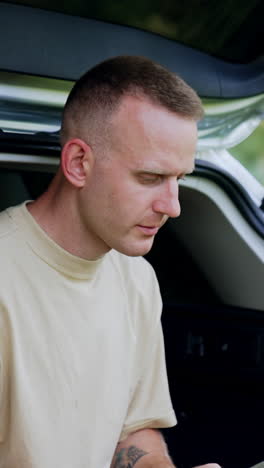 focused male freelancer looks at the screen of his laptop. man works remote sitting in the car trunk in forest. vertical video