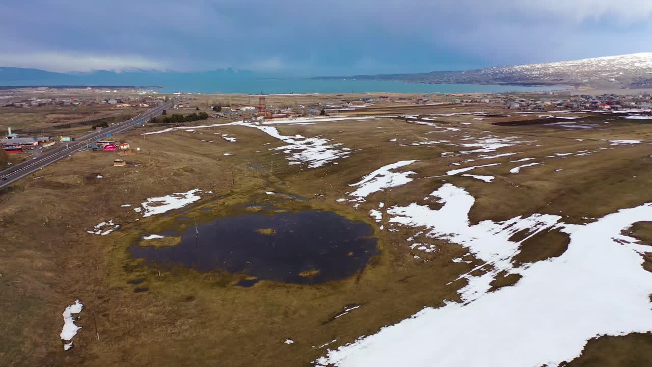 Landsby nær havet med motorvej og spredte huse, droneudsigt over vinterlandskab, pletvis sne, overskyet himmel, fjerne bjerge.